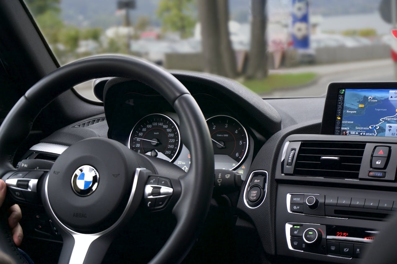 Interiorul unui automobil BMW, cu volanul, tabloul de bord și sistemul de navigație vizibile. Se observă logo-ul BMW pe volan și diferite comenzi și butoane pe consola centrală. Pe fundal, printr-o fereastră, se pot vedea clădiri și un peisaj urban