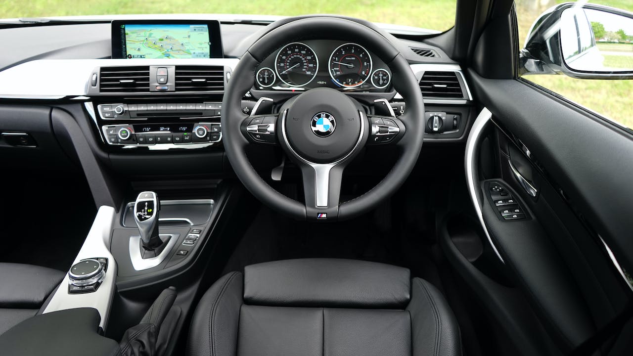 Interiorul unui automobil BMW, cu volan cu logo BMW în centru, ecran de navigație luminat, schimbător de viteze automat și detalii de design sofisticate. Scaunele din piele de culoare neagră și detalii argintii pe consolă subliniază luxul și stilul modern al mașinii