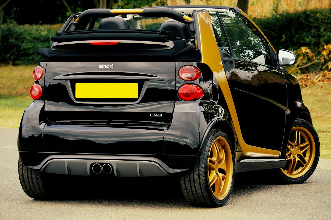 Vizualizare din spate a unui automobil "Smart" negru cu accente galbene, echipat cu o etichetă "Brabus", și cu roți aurii, parcat lângă copaci